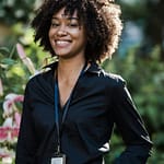 A confident woman with an ID card smiles outdoors in a lush garden.