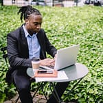 Concentrated African American businesswoman in earbuds wearing formal black suit sitting at table in city garden and typing on netbook