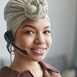 Delighted African American female call agent with headset wearing turban looking away while standing in light room on blurred background