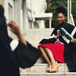 Black female graduate in gown smiles with diploma on city stairs, embodying achievement.