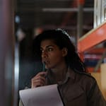 A woman in a warehouse contemplatively checking inventory under low light conditions.
