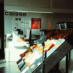 Grocery store interior with neatly arranged fruit display and promotional poster at checkout area.