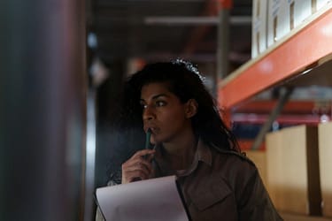 A woman in a warehouse contemplatively checking inventory under low light conditions.