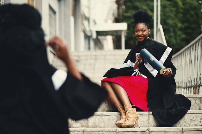 Black female graduate in gown smiles with diploma on city stairs, embodying achievement.